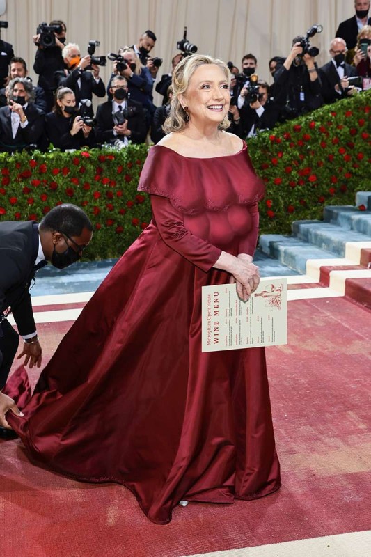 ILLUSIONS OF GRANDEUR — The MTE poses for press cameras as her “footman” stoops to retrieve her dropped colostomy bag at a MET gala in Manhattan, May 2, 2022.